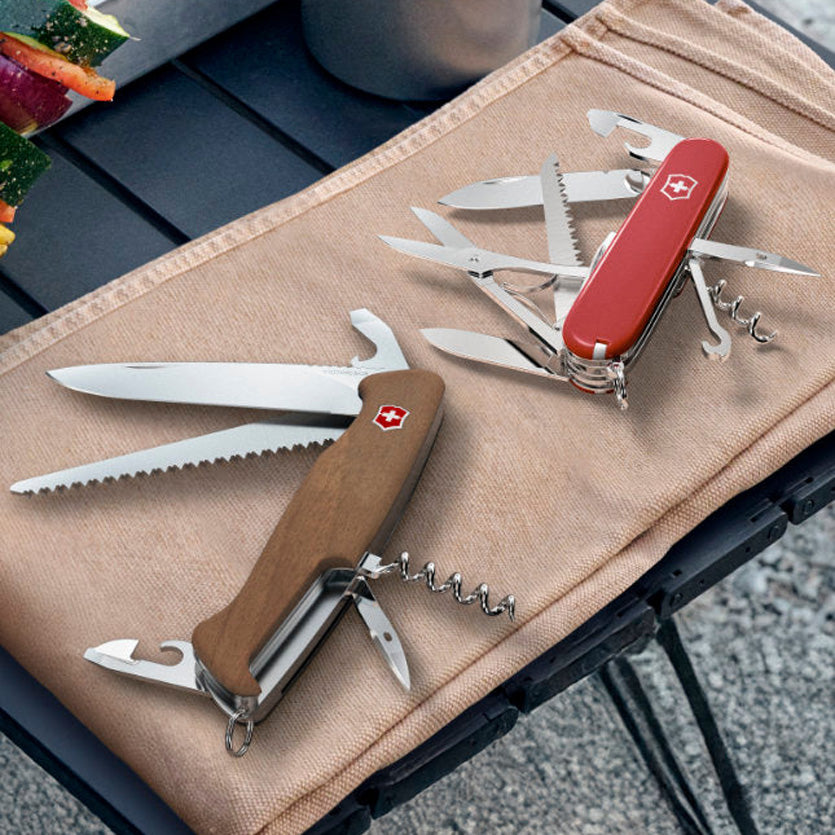 Two Swiss Army knives on a beige cloth with a blurred outdoor background